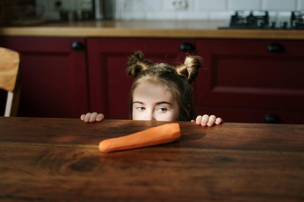 Cute child with buns peeks at a fresh carrot on a rustic kitchen table.