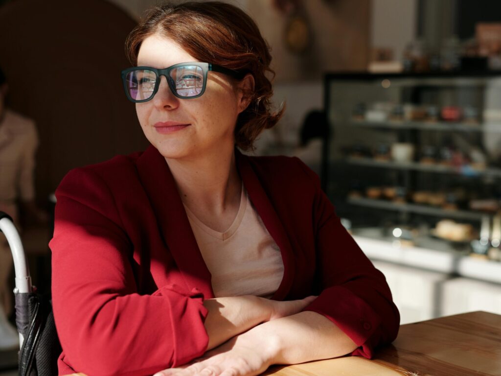 Woman in a wheelchair enjoying a sunny day at a café, wearing eyeglasses and a red jacket.