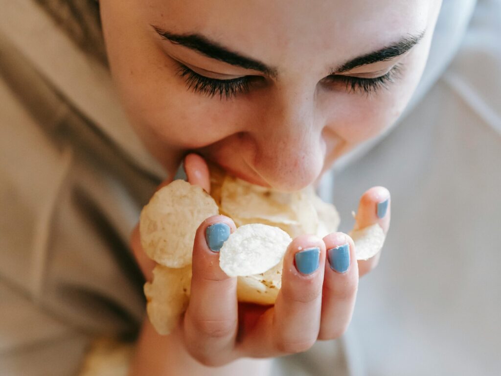 From above of crop female in sweater with black hair and handful of potato chips eating unhealthy fast food on blurred background