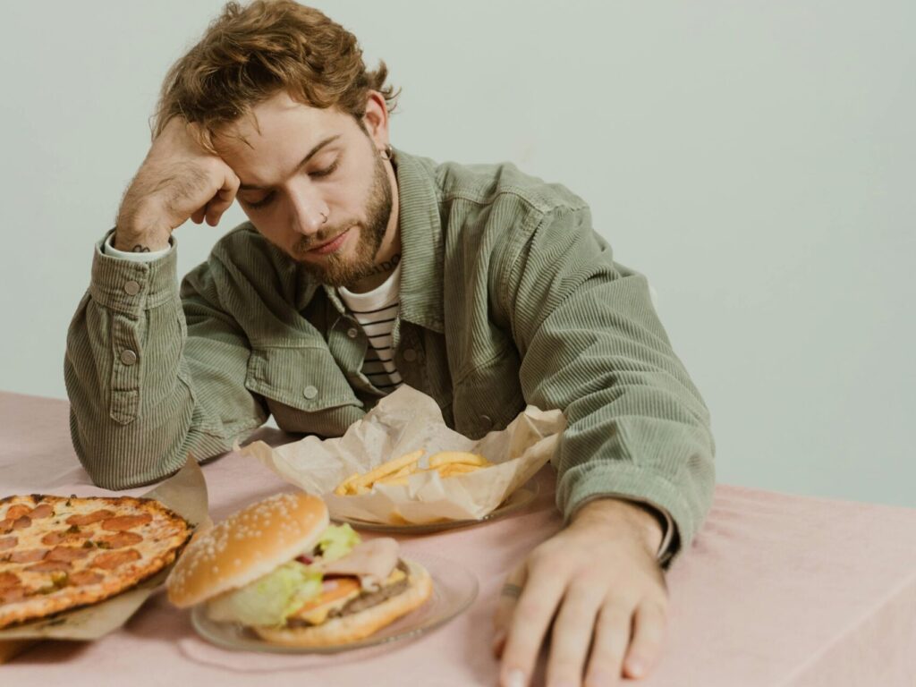 Young man appears tired with fast food burger and pizza on table in a relaxed setting.