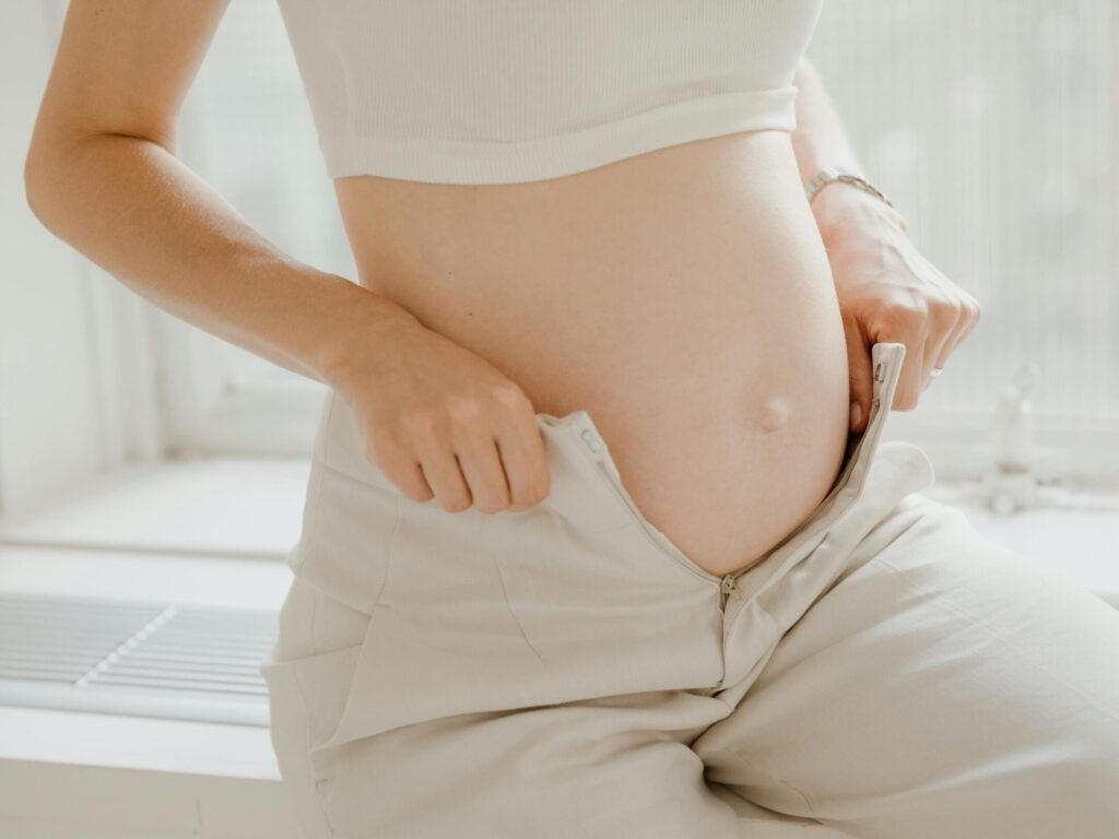 A close-up of a pregnant woman adjusting her pants, highlighting her belly in a sunlit room.